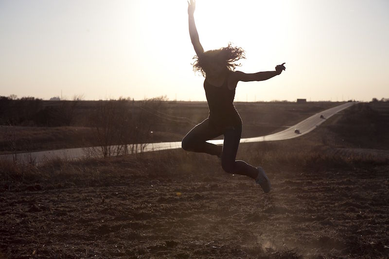 A woman in a field on the side of a highway leaps into the air as the sun sets
